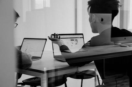 Two individuals are engaged in a discussion while working on laptops in a modern office setting.
