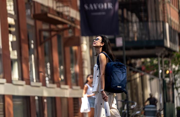 A woman wearing sunglasses and a backpack walks down the street, looking up at the buildings.