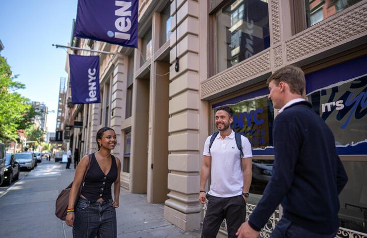 Three young adults are chatting on a sunny street in front of a building with banners.