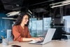 A woman in a polka dot shirt smiling and working on a laptop in a modern office environment.