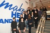 A group of young people and adults poses on a staircase in front of a large wall sign that reads 'make it HER and then'.