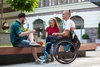 Three friends, one in a wheelchair, chatting and smiling in an outdoor urban setting.