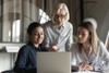 An older woman instructs two younger women using a laptop in an office setting.