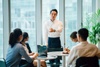 A man stands presenting to colleagues at a round table in a brightly lit office.