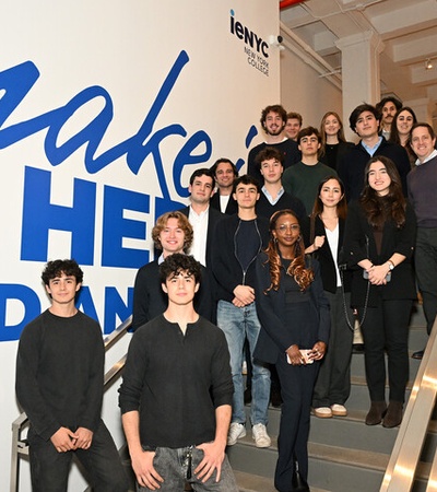 A group of young people and adults poses on a staircase in front of a large wall sign that reads 'make it HER and then'.