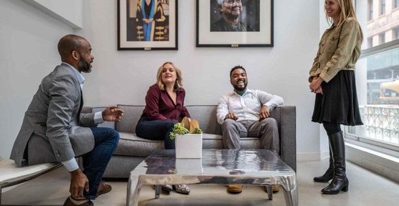 Four people, two men and two women, are having a lively conversation in a modern office lounge area.