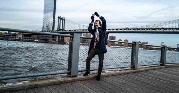A person is taking a picture with a smartphone by a river with a bridge in the background.