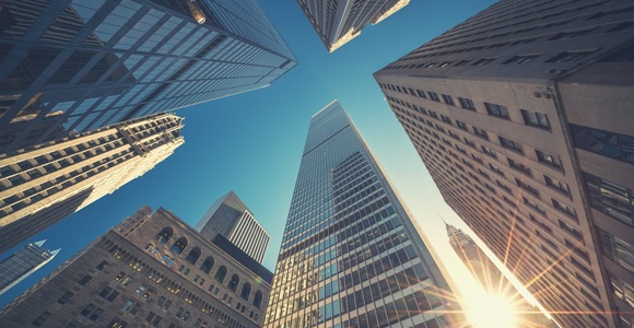 Looking up at towering skyscrapers against a clear blue sky with sunlight shining through.
