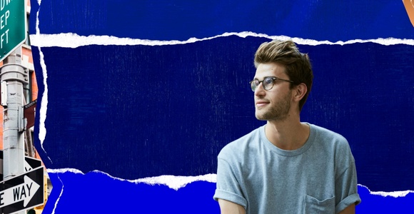 A young man wearing glasses is sitting against a vibrant blue background with street signs in the foreground.