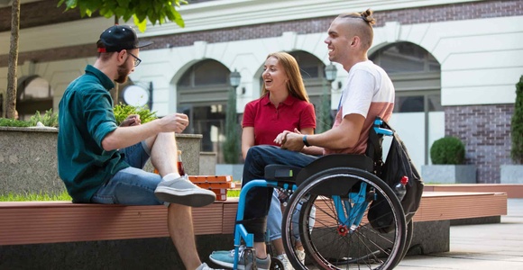 Three friends, one in a wheelchair, chatting and smiling in an outdoor urban setting.